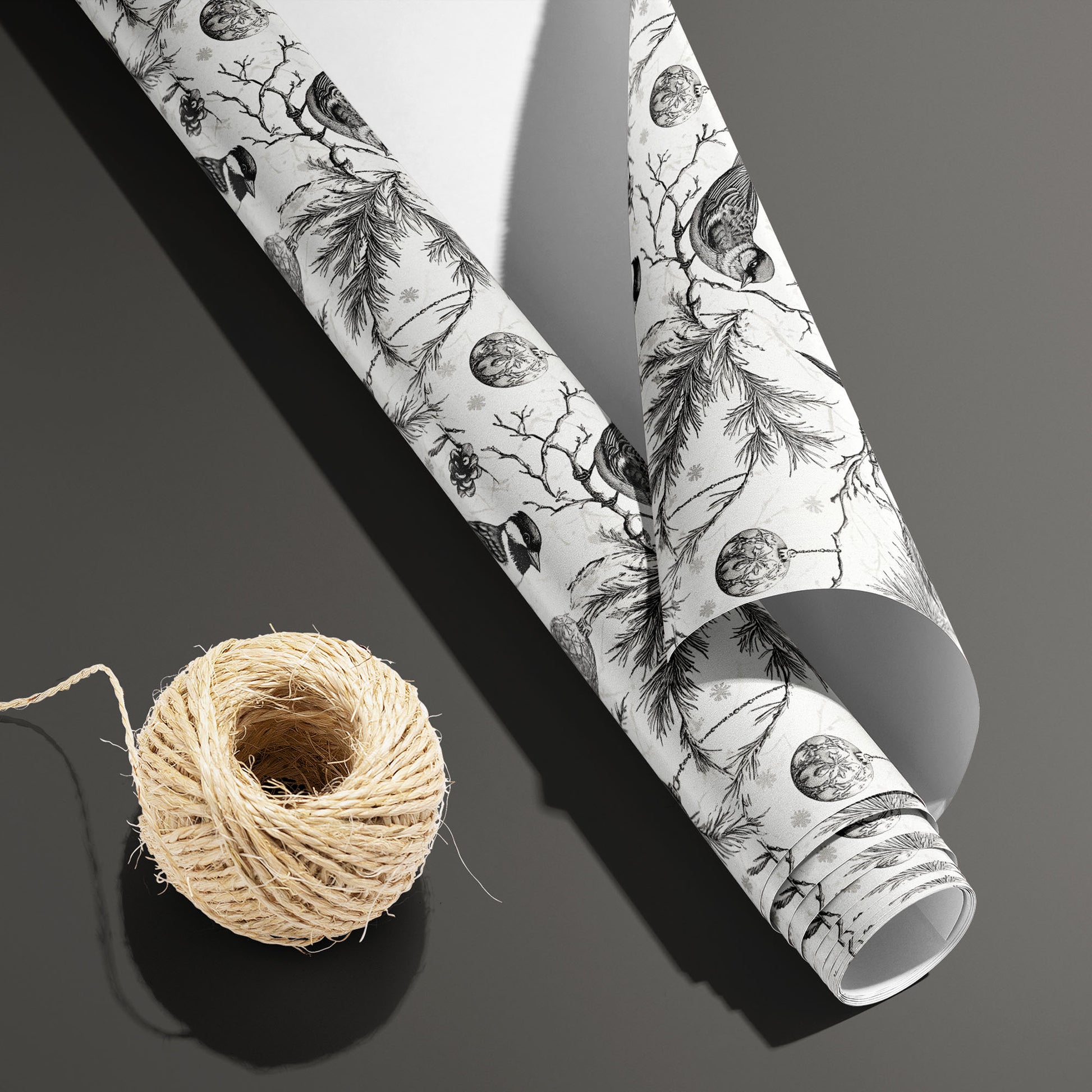 A partially unfurled Ornamental Nesting roll with its white underside beside a spool of natural twine on dark gray, displaying birds, pine boughs, cones, and ornaments on white.
