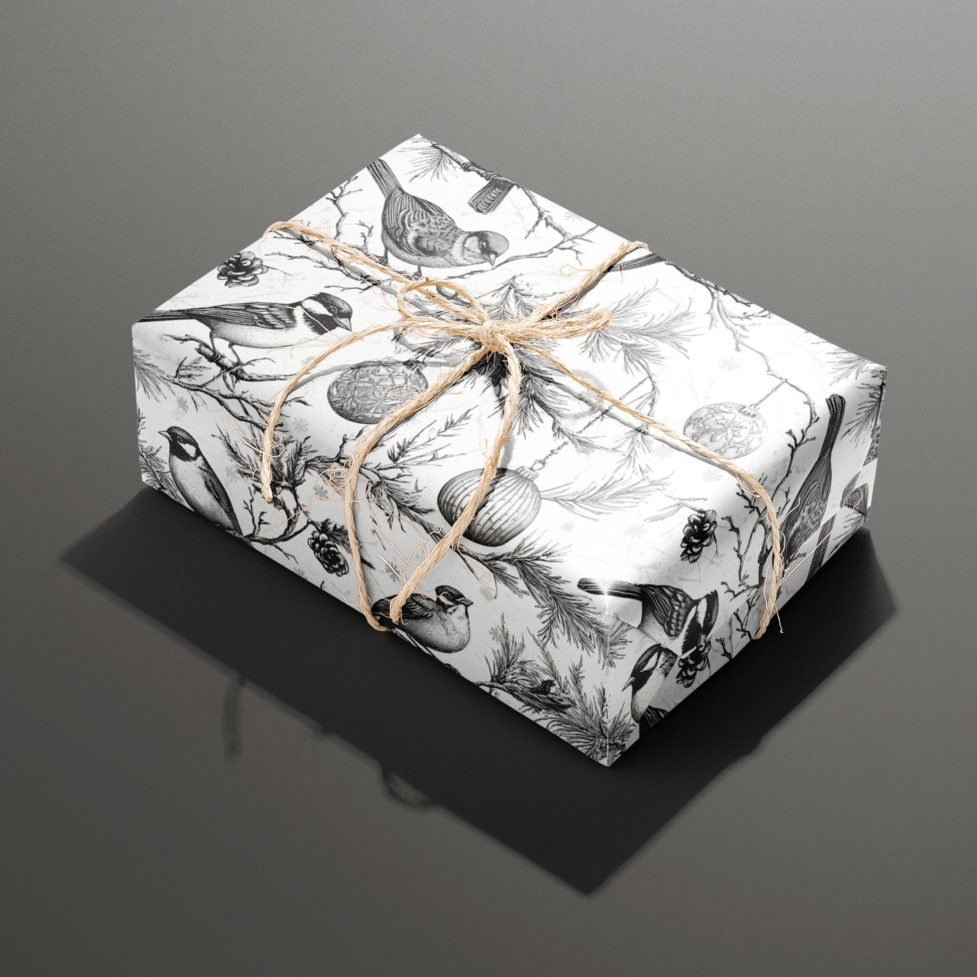 A single rectangular gift wrapped in Ornamental Nesting and tied with natural twine, the monochrome birds, ornaments, and pine boughs crisp on a white background over dark gray.