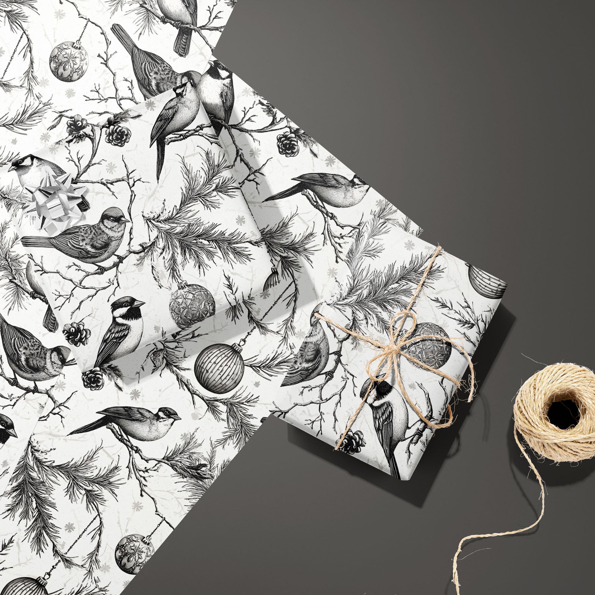 Two gifts in Ornamental Nesting—one with a silver bow, one tied with twine—rest on two unused sheets beside a twine spool on a dark gray surface, with birds and ornaments.