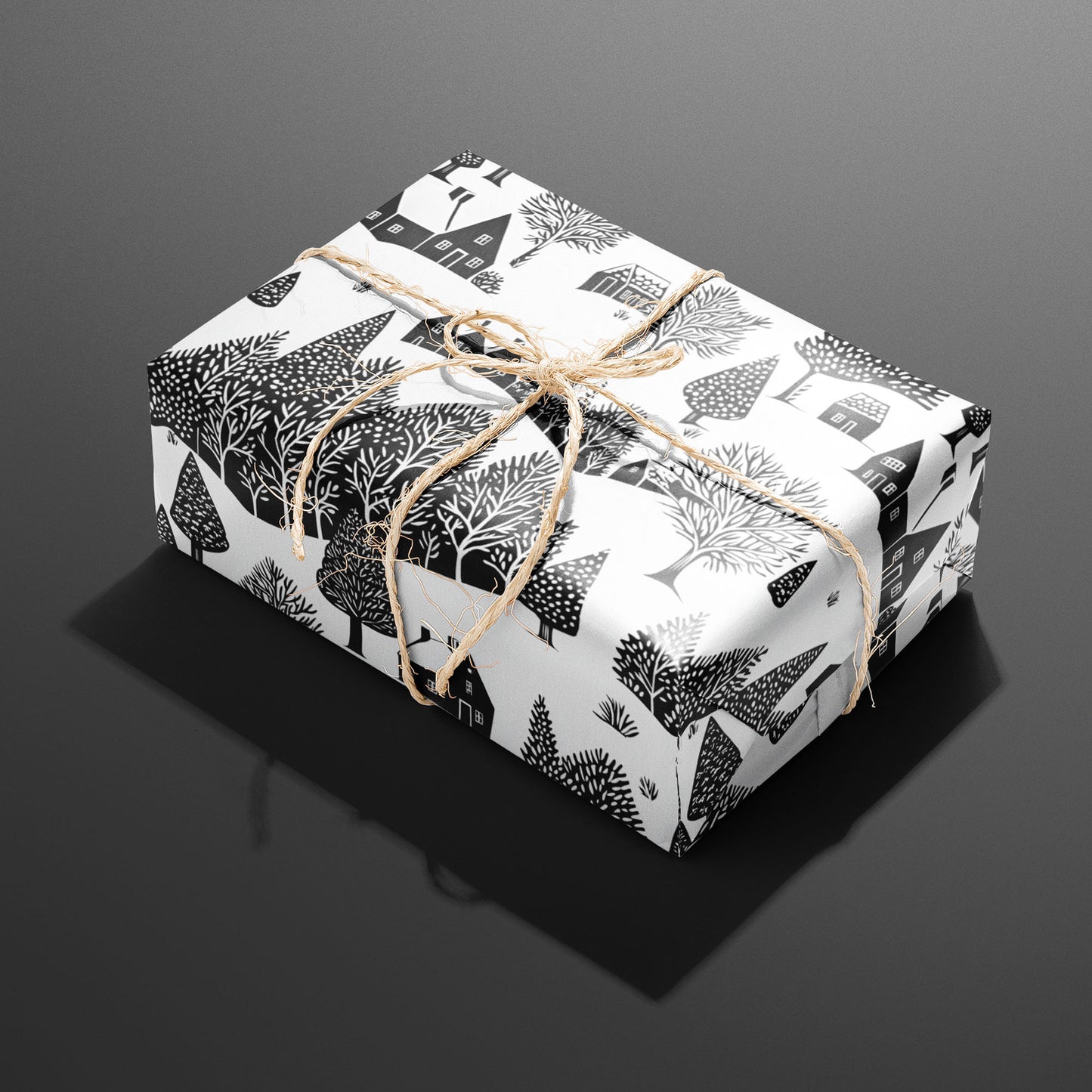 A single rectangular gift wrapped in Snowbound Village and tied with natural twine, the black cottages and snow-laden trees crisp on a white ground over dark gray.