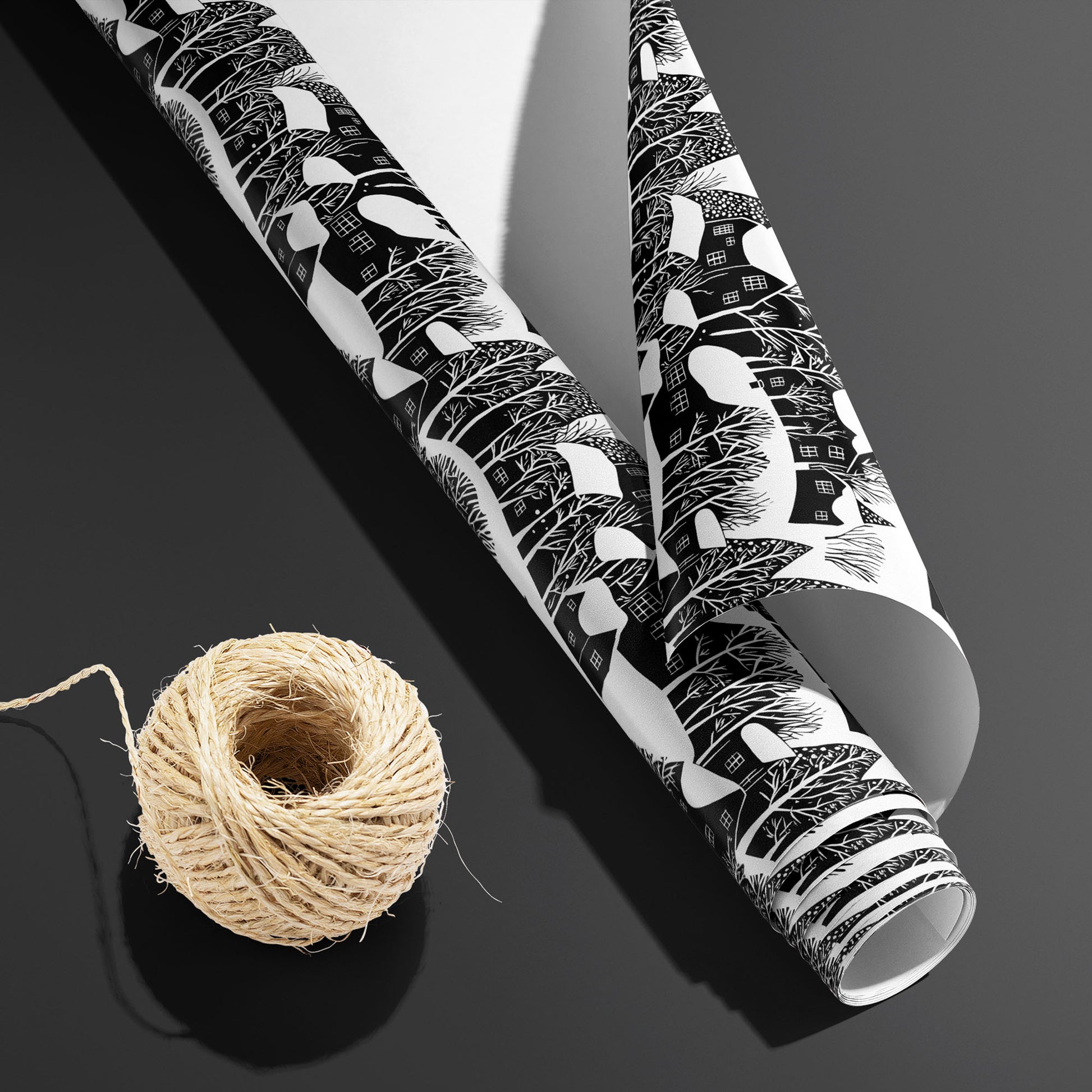 A partially unfurled roll of Snowy Silhouette, white underside visible next to a spool of natural twine, shows winter village silhouettes on a charcoal gray surface.
