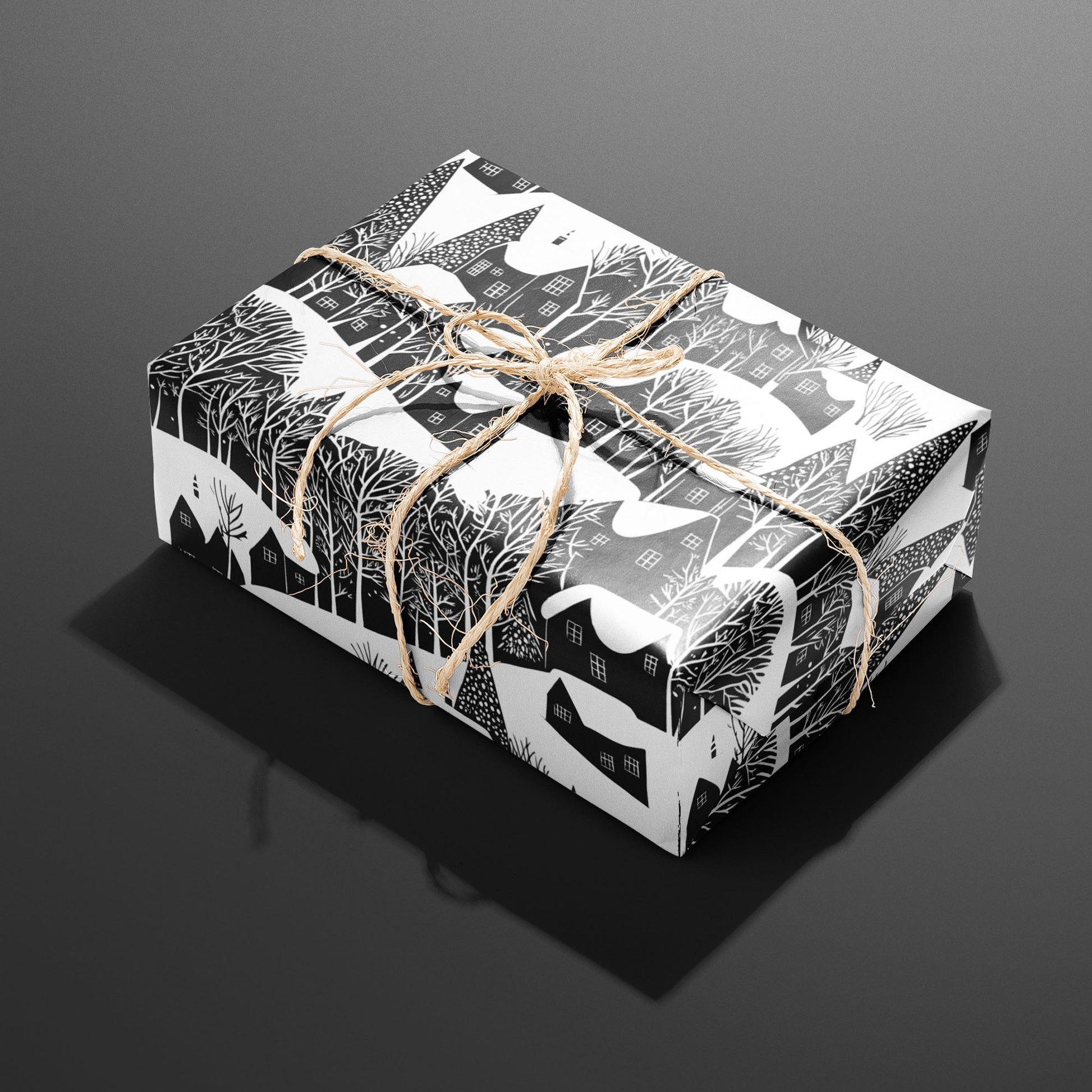 A single rectangular gift wrapped in Snowy Silhouette and tied with natural twine highlights snowy houses and leafless trees in black and white on a charcoal gray backdrop.