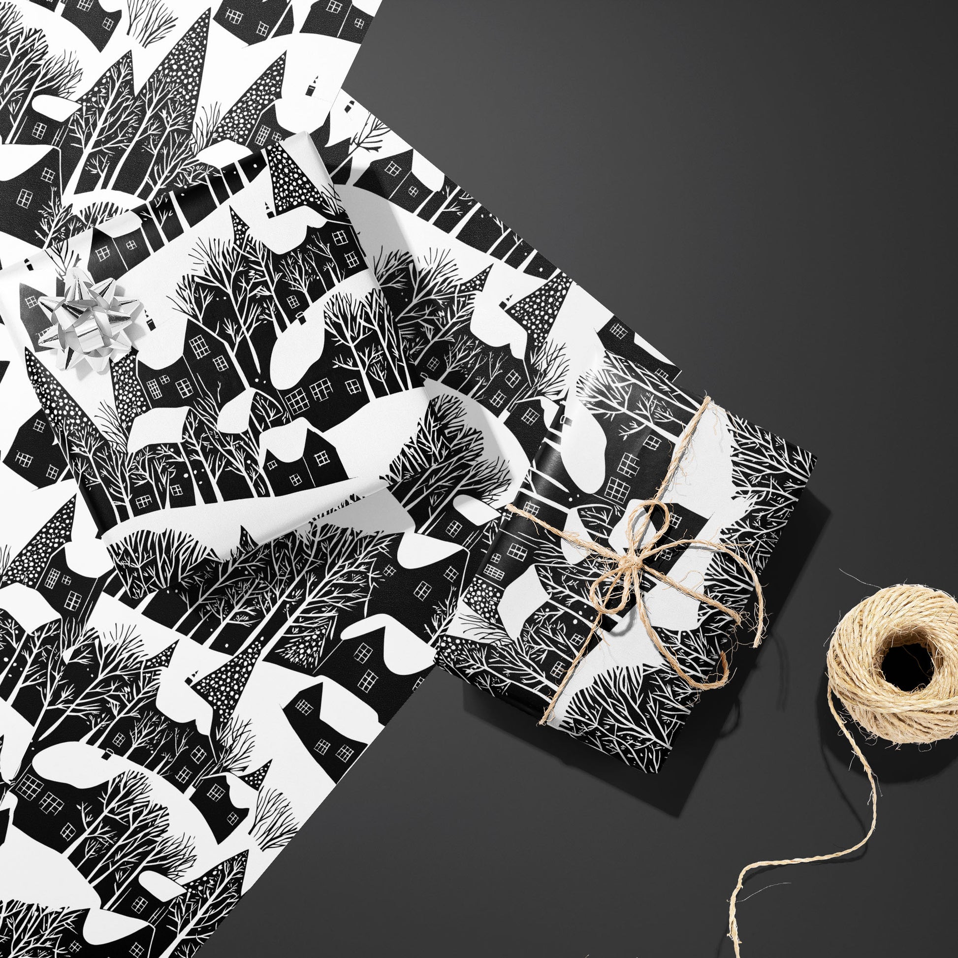 Two Snowy Silhouette gifts—one with a silver bow, one tied with twine—rest on two unused sheets beside a twine spool on a charcoal gray surface, showing snowy village silhouettes.