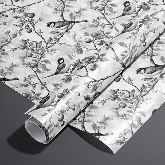 A roll on an unrolled sheet of Winter Orchard wrapping paper with etched songbirds on fruiting branches over a frosty gray ground, set on a dark charcoal background.
