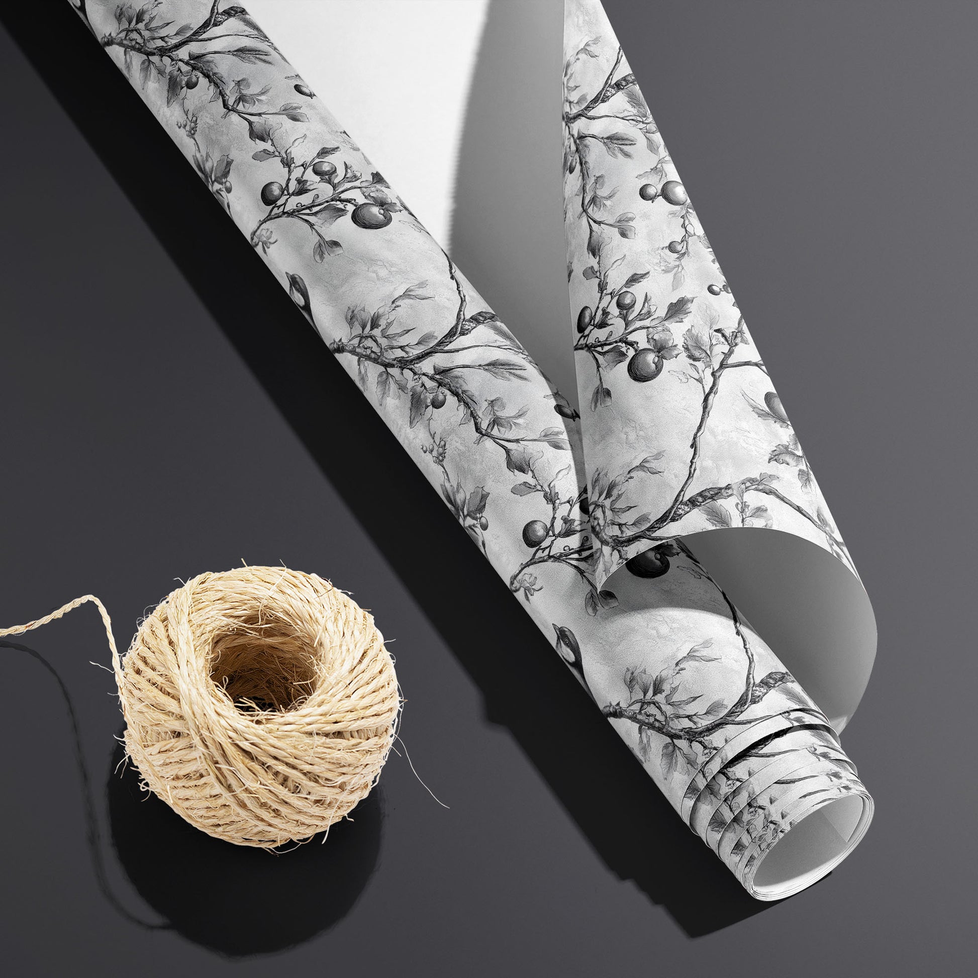 A partially unfurled Winter Orchard roll with the white underside visible beside a spool of natural twine, featuring etched birds and fruiting branches on a dark charcoal background.