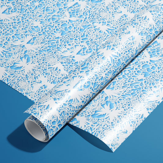 A roll of Winter's Whisper rests on a matching unrolled sheet, showing white lace-cut winter florals and snowflakes over icy blue on a deep blue surface.
