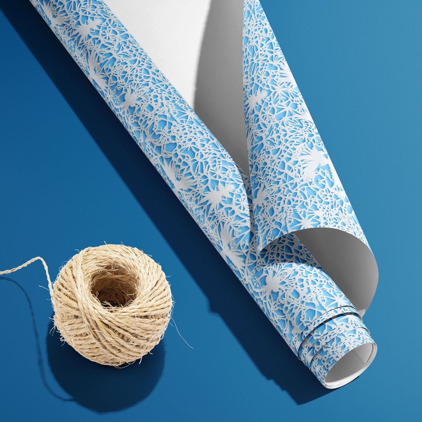 A partially unfurled roll of Winter's Whisper, white underside visible beside natural twine, displays lace-like winter florals and snowflakes on icy blue on a deep blue surface.