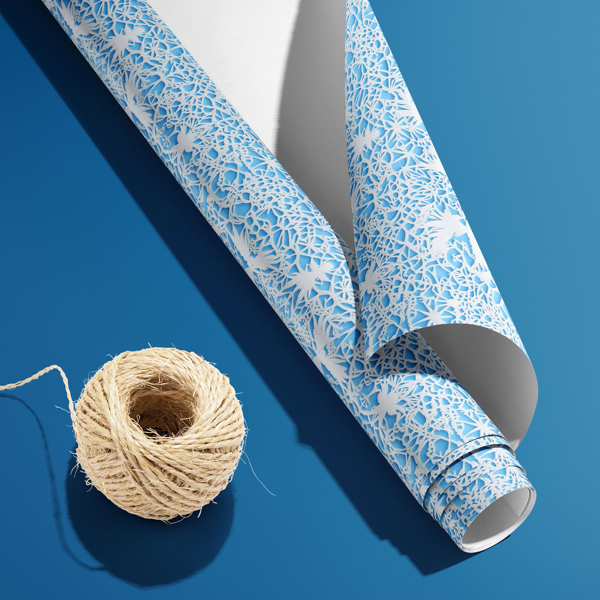A partially unfurled roll of Winter's Whisper, white underside visible beside natural twine, displays lace-like winter florals and snowflakes on icy blue on a deep blue surface.
