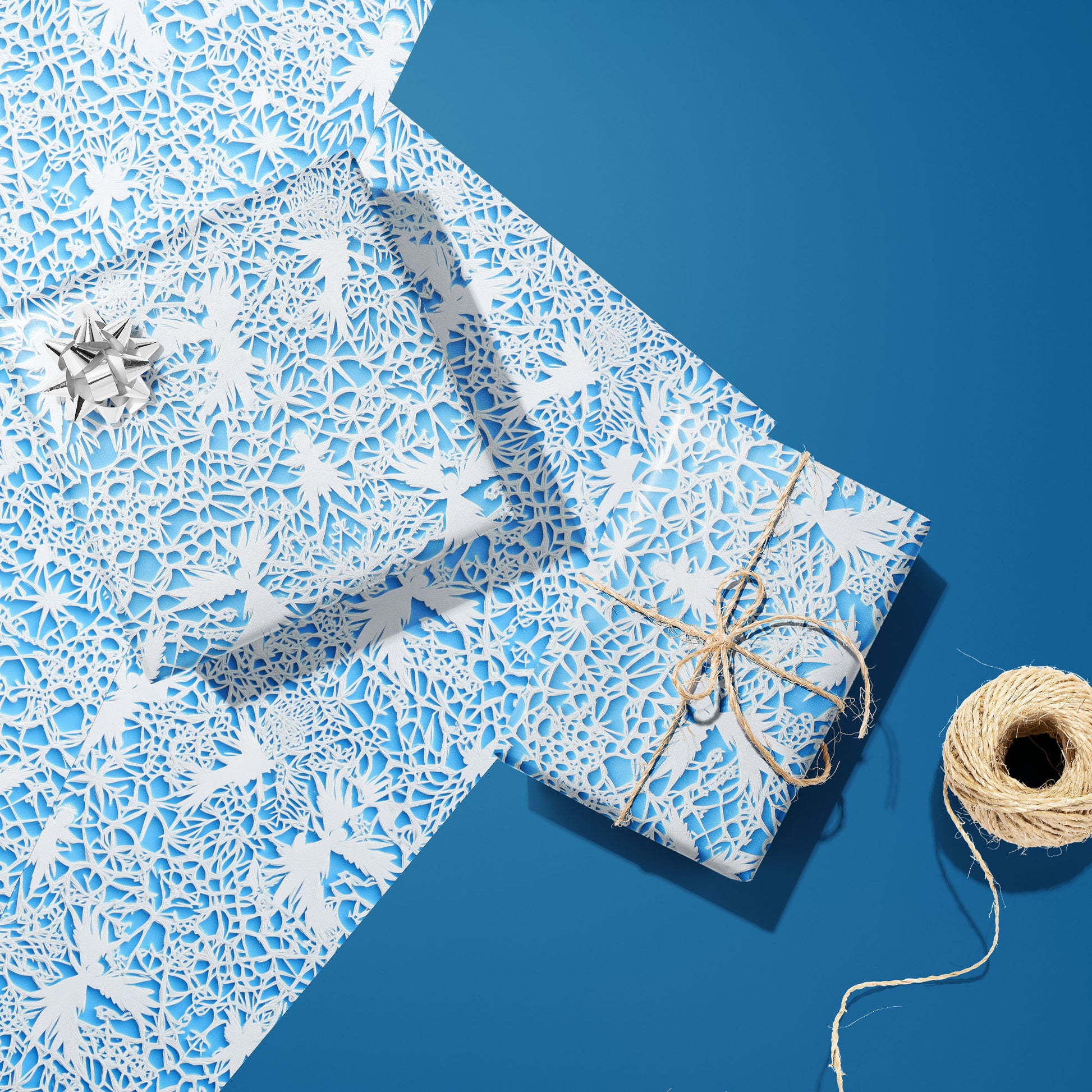 Two gifts in Winter's Whisper—one with a silver bow and one tied with twine—sit on two unused sheets with white lace winter florals on icy blue, on a deep blue surface beside twine.