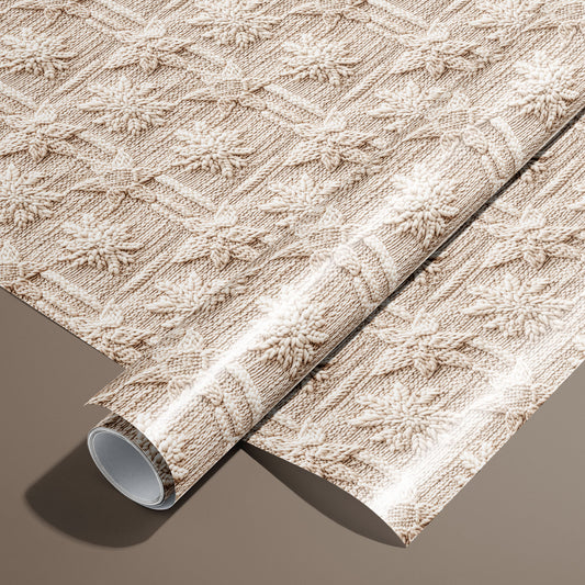 A roll of Winter Weave wrapping paper rests on an unrolled sheet, showing a cozy knit-style snowflake pattern in cream and beige on a warm brown surface.