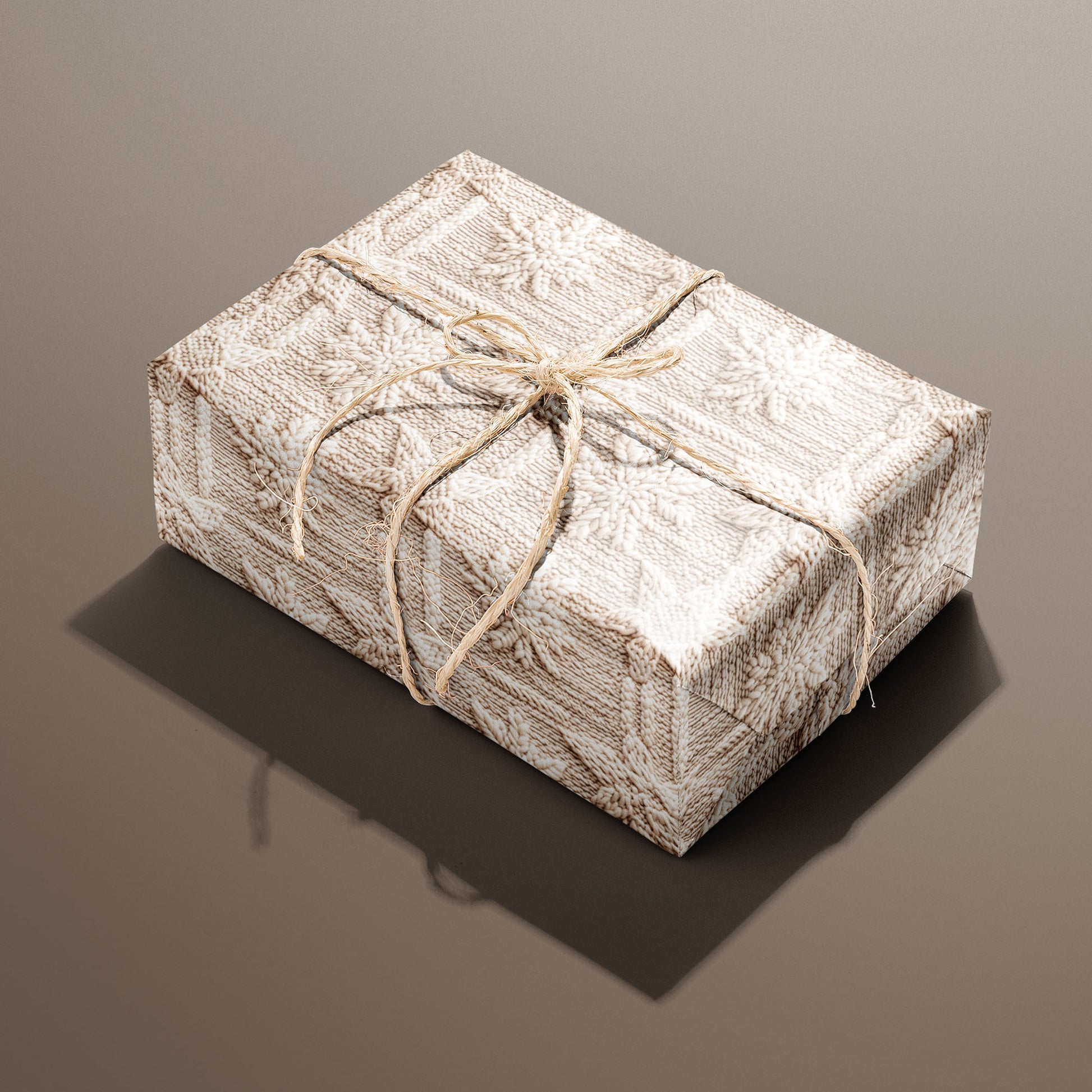 A single rectangular gift wrapped in Winter Weave and tied with natural twine highlights a cozy knit snowflake design in cream and beige on a warm brown backdrop.
