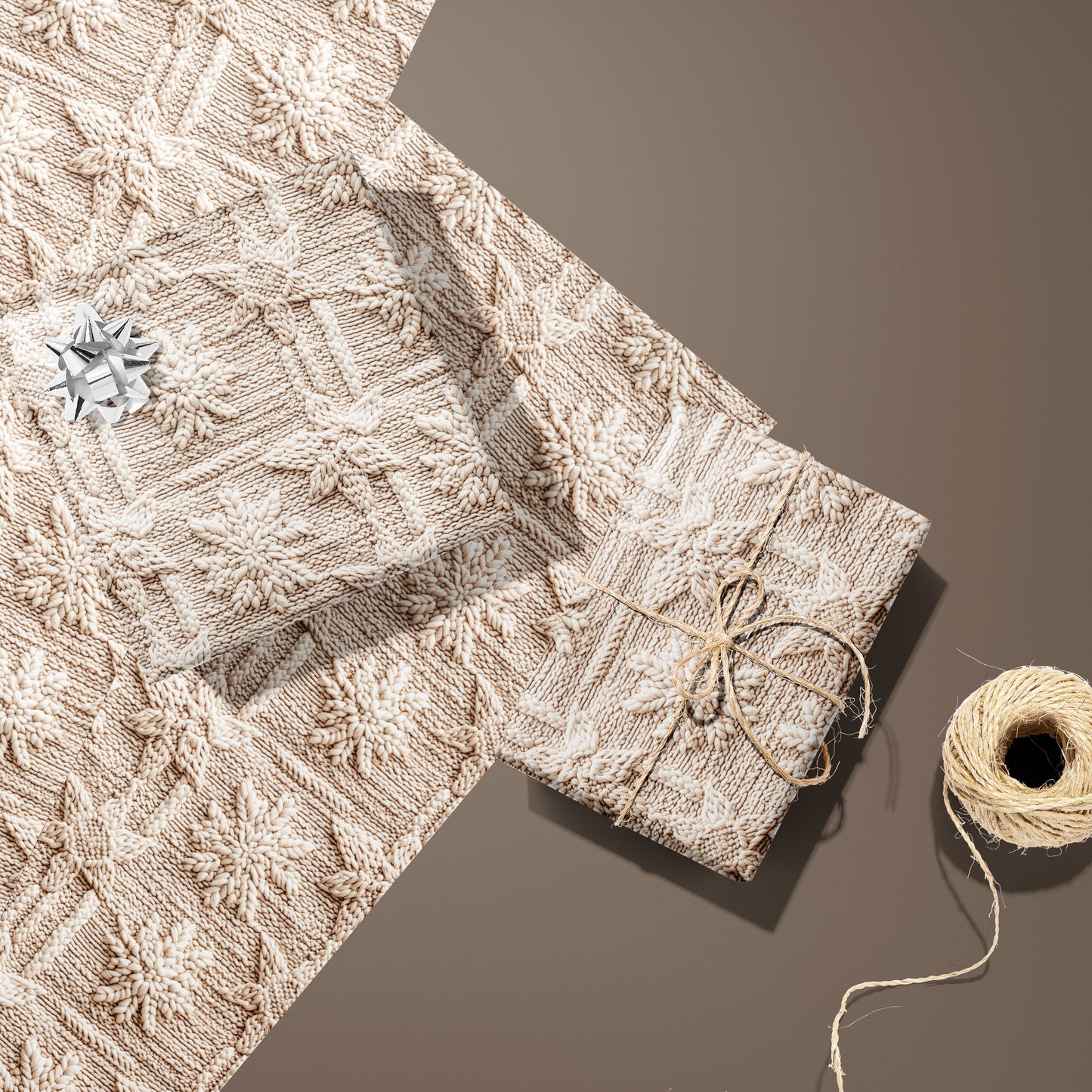 Two gifts in Winter Weave—one with a silver bow, one tied with twine—rest on two unused sheets beside a twine spool on a warm brown surface, showing a knit snowflake pattern.