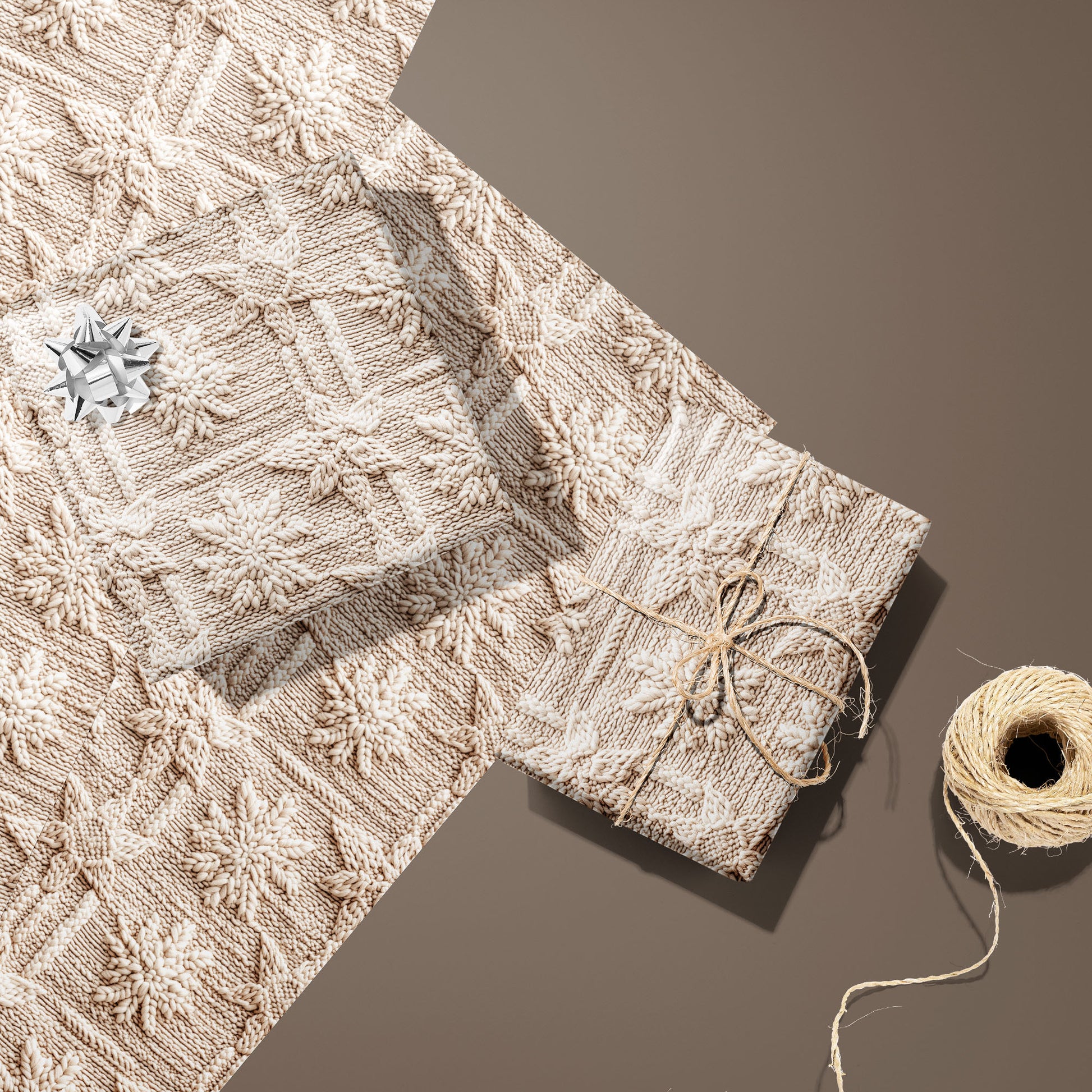 Two gifts in Winter Weave—one with a silver bow, one tied with twine—rest on two unused sheets beside a twine spool on a warm brown surface, showing a knit snowflake pattern.