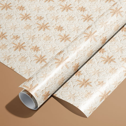 A roll of Hygge Holiday wrapping paper resting on a matching unrolled sheet, featuring knit-texture Scandinavian snowflakes in warm tan on soft white atop a caramel-brown surface.
