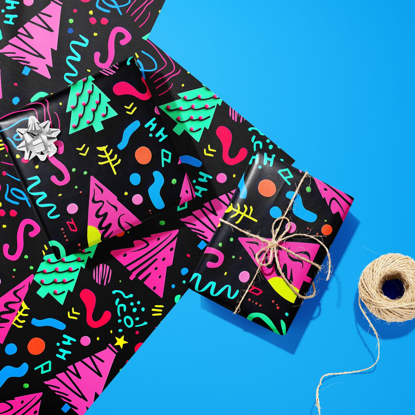 Two gifts wrapped in Electric Wonderland—one with a silver bow and one tied with twine—rest on two unused sheets beside a twine spool on bright blue, showing neon Memphis trees and squiggles.