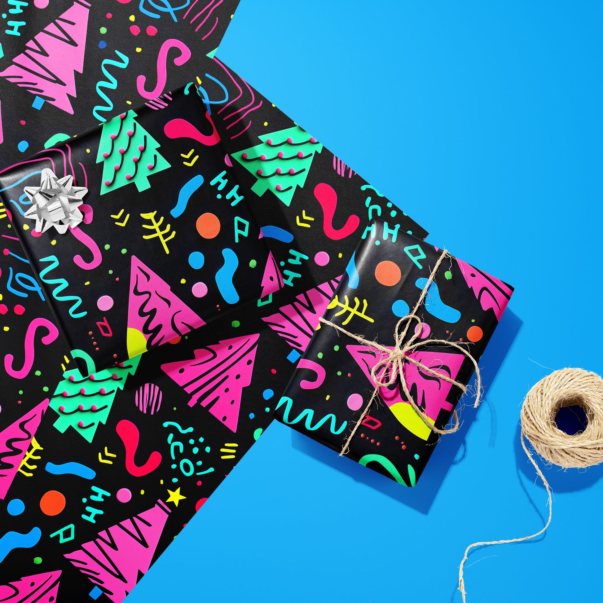 Two gifts wrapped in Electric Wonderland—one with a silver bow and one tied with twine—rest on two unused sheets beside a twine spool on bright blue, showing neon Memphis trees and squiggles.