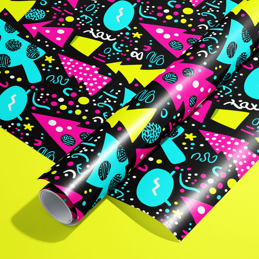 A roll on an unrolled sheet of Neon Noel wrapping paper with Memphis-style trees, ornaments, squiggles, and dots in magenta, cyan, and chartreuse on black, set on an acid-yellow background.
