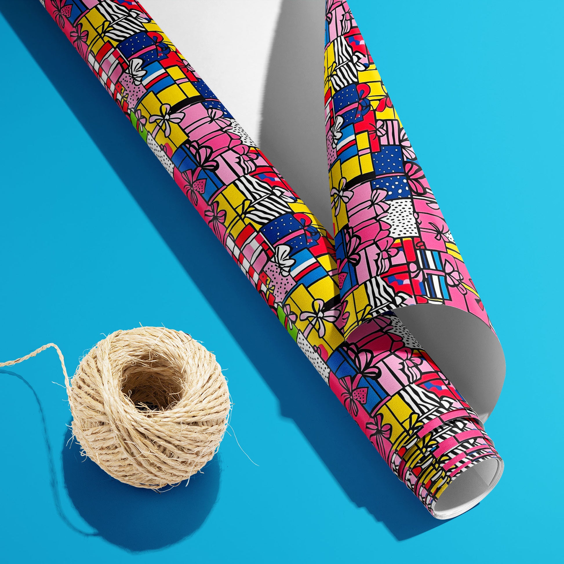 A partially unfurled roll of Pop Art Presents, white underside visible beside a spool of natural twine, shows pop-art presents in bold colors on a bright cyan surface.