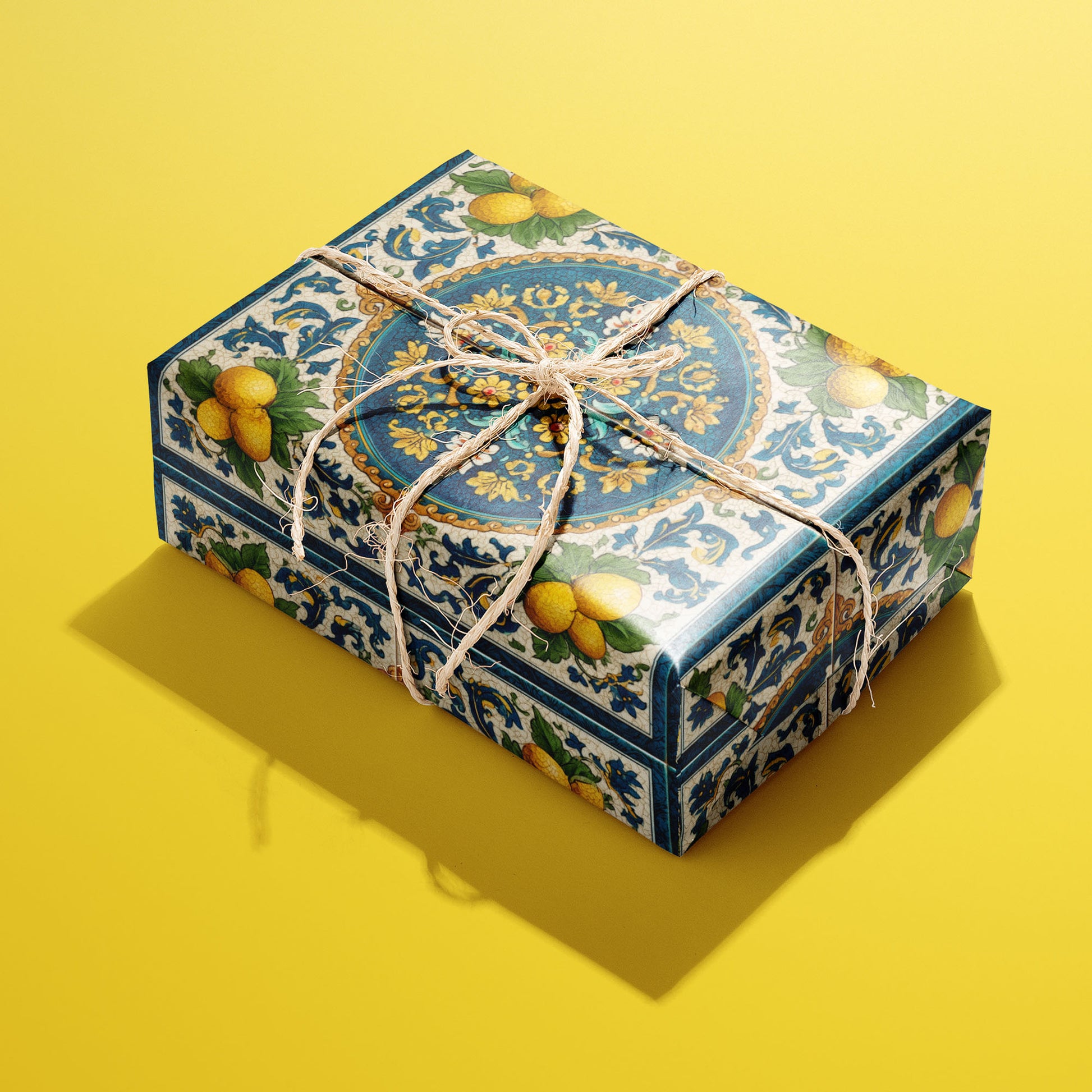 A rectangular gift wrapped in Citron Azulejo paper, the lemon-and-azulejo tile design tied with a natural twine bow, set on a bold yellow background.