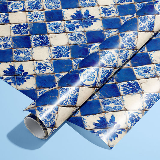 A roll of Ionian Inks wrapping paper resting on a full sheet, featuring cobalt-and-ivory azulejo checker tiles with floral motifs on a pale blue background.