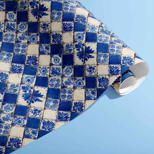 A partially unfurled roll of Ionian Inks wrapping paper showcasing blue-and-white ceramic tile florals in a rustic checker pattern against a light blue surface.