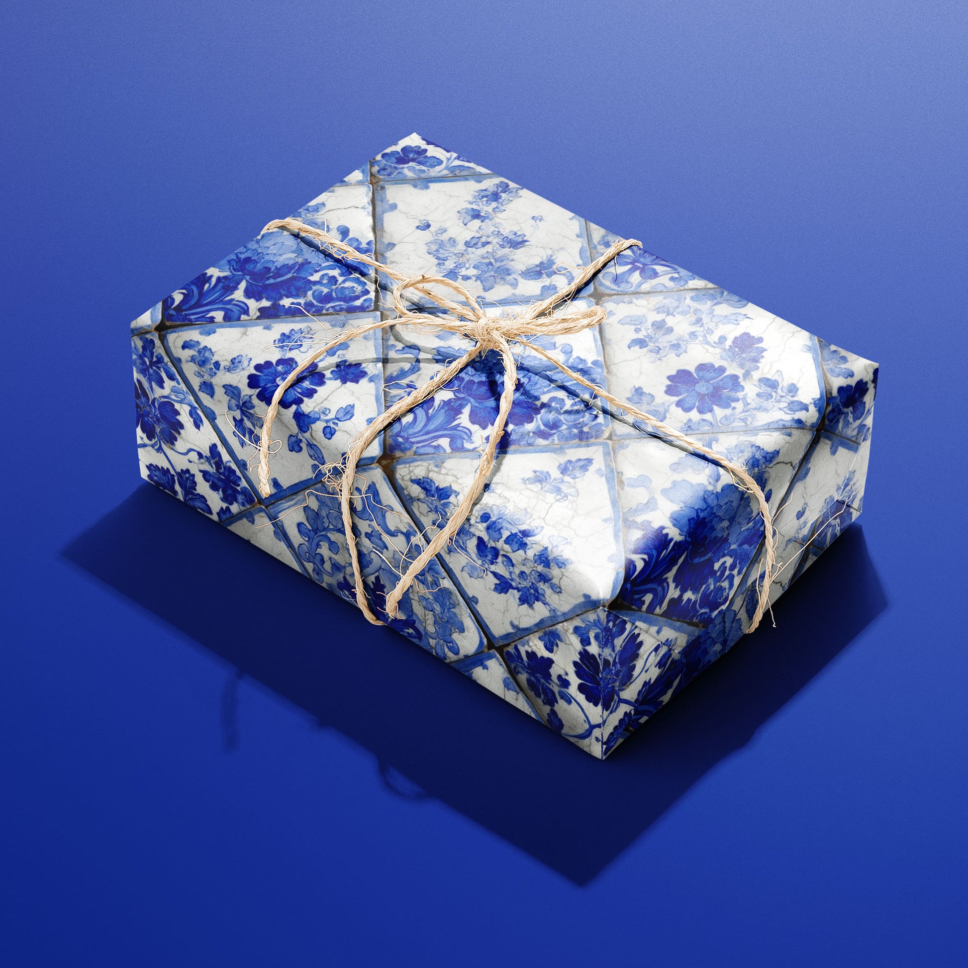 A rectangular gift wrapped in Aegean Bouquet paper, the blue-and-white tile florals tied with a natural twine bow, set on a saturated royal blue background.