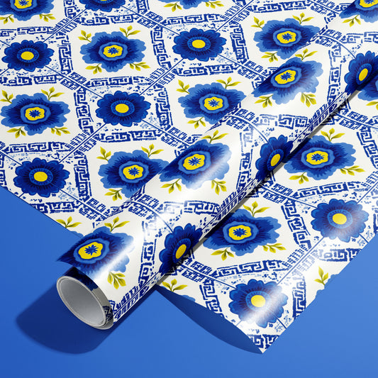 A roll of Santorini Floriate wrapping paper resting on an unrolled sheet, featuring cobalt floral tile motifs with Greek-key lines on white, set against a bold blue background.