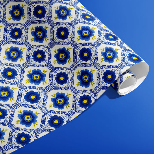 A partially unfurled roll of Santorini Floriate gift wrap showing blue blossom-and-tile motifs on white, displayed diagonally over a vivid blue backdrop.