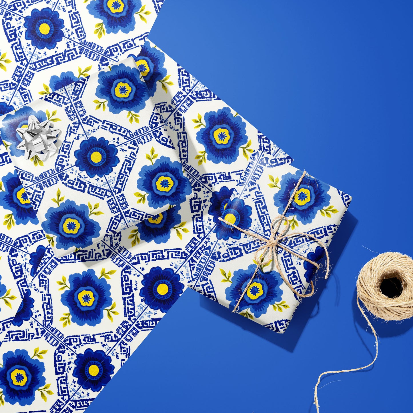 Two gifts wrapped in Santorini Floriate—one with a silver bow, one tied with twine—placed on two unused sheets of the blue floral tile print beside a twine spool on a blue surface.