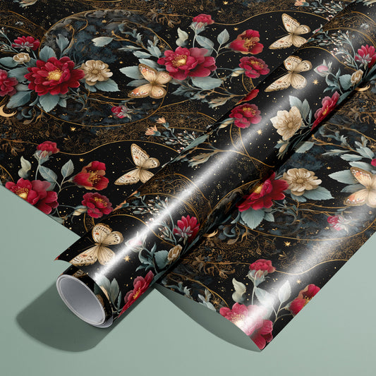 A roll resting on an unrolled sheet of Velvet Asterism wrapping paper, a black celestial floral with red and cream blooms, butterflies, and gold filigree, on a sage green surface.