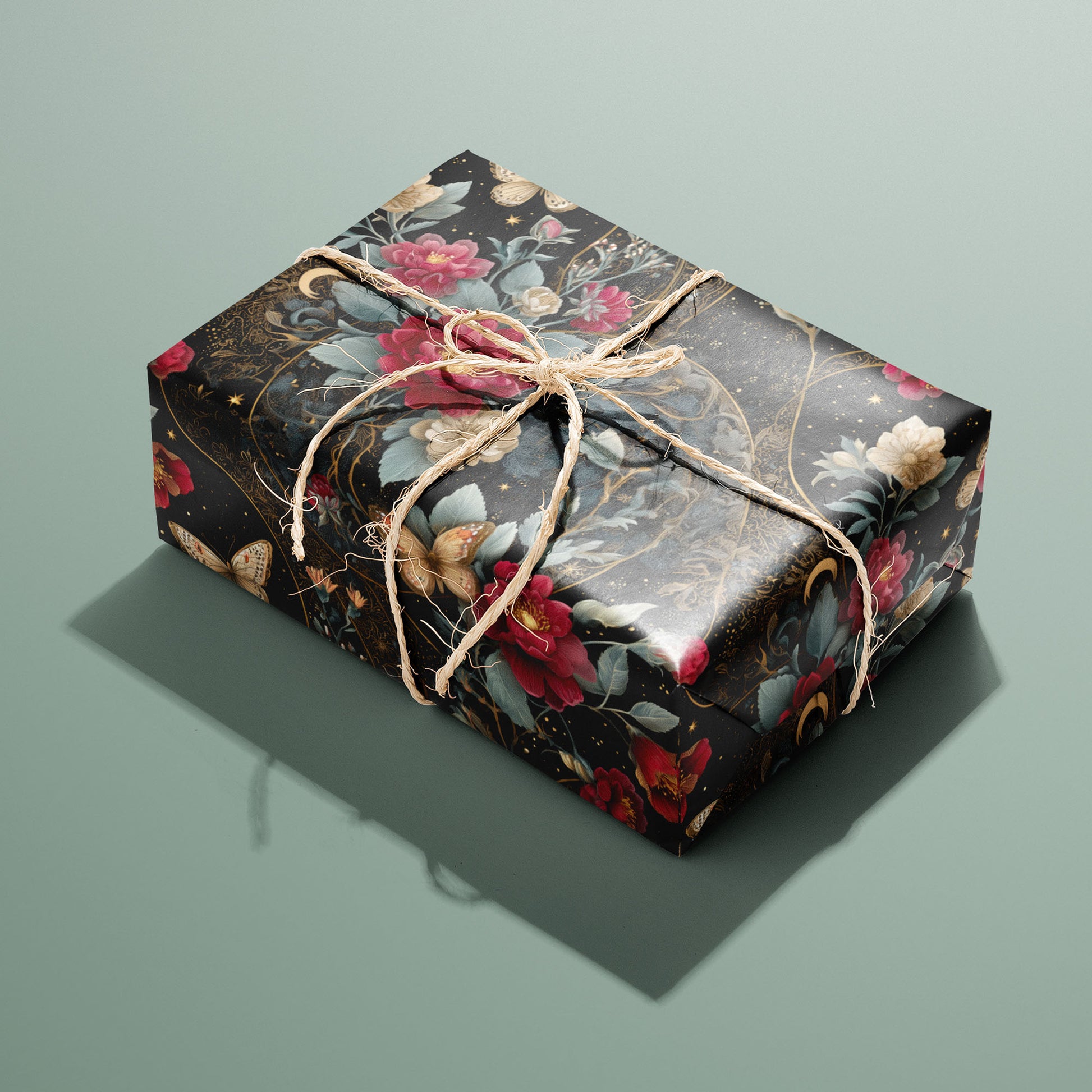 A single gift wrapped in Velvet Asterism paper—black celestial florals with butterflies and gold filigree—tied with natural twine on a sage green tabletop.