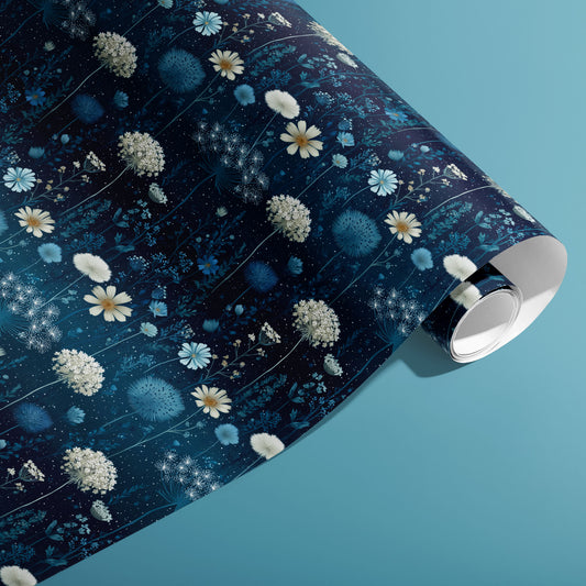 A partially unfurled roll of Midnight Garden wrapping paper showing blue botanical blooms and tiny starry dots on a deep navy ground, on a teal background.