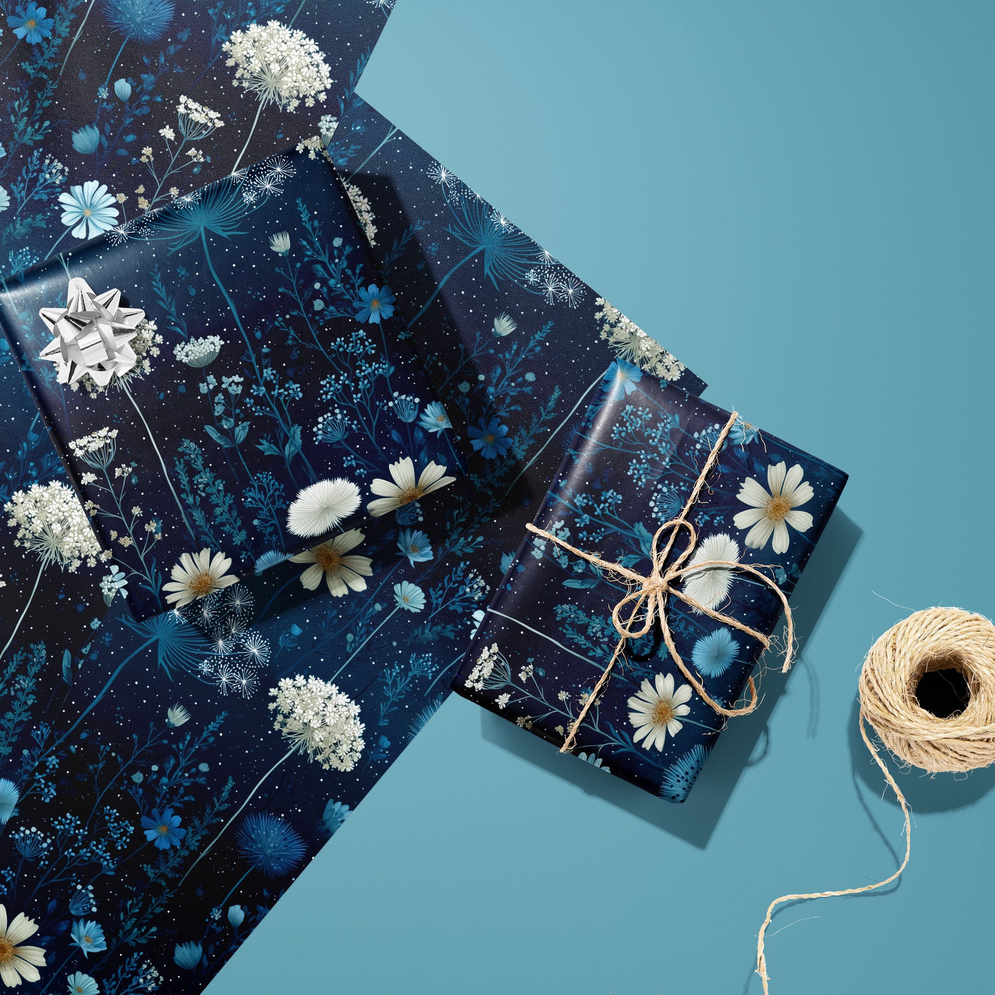 Two gifts in Midnight Garden wrap—one with a silver bow, one tied with twine—rest on two unused sheets beside a spool of twine on a teal background.