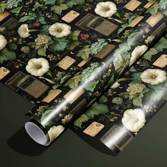 A roll of Vesper Botanica wrapping paper on an unrolled sheet, featuring vintage florals, apothecary labels, and tiny moons on black, set on a dark olive green surface.