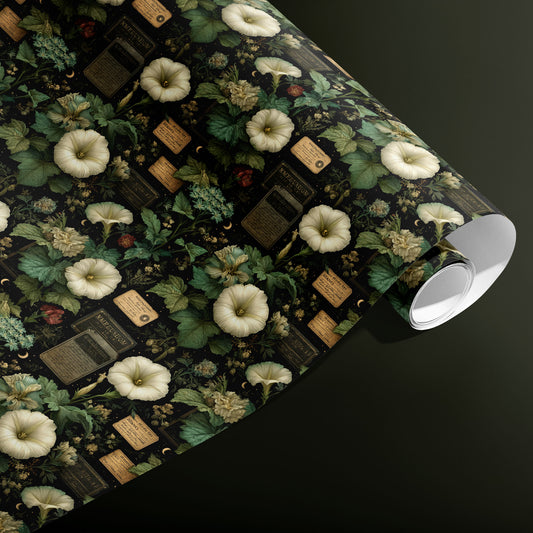 A partially unfurled roll of Vesper Botanica wrapping paper showing cream blossoms, lush leaves, and antique labels with celestial accents on black, on a dark olive green surface.