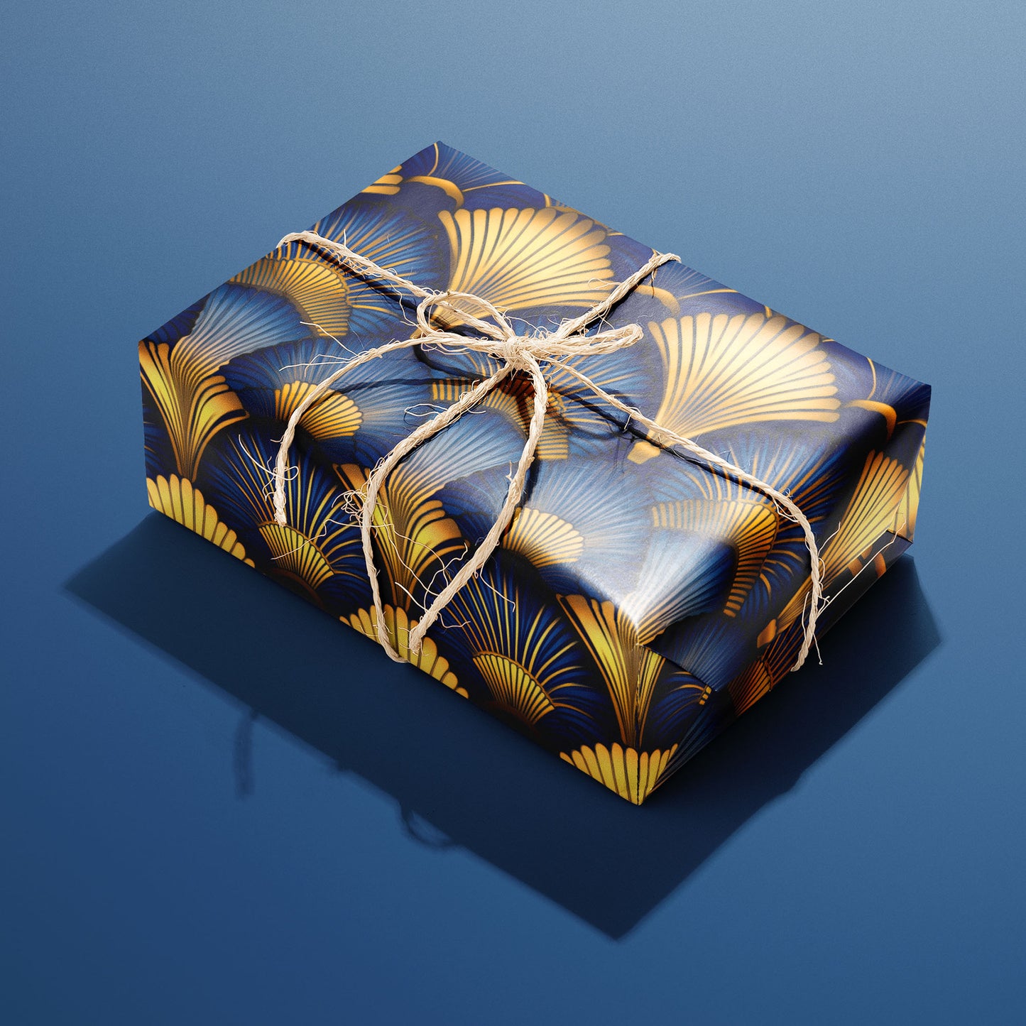 A single gift wrapped in Gilded Ginko and tied with natural twine displays gold and blue ginkgo leaves and delicate stems on a dark field, sitting on a blue background.