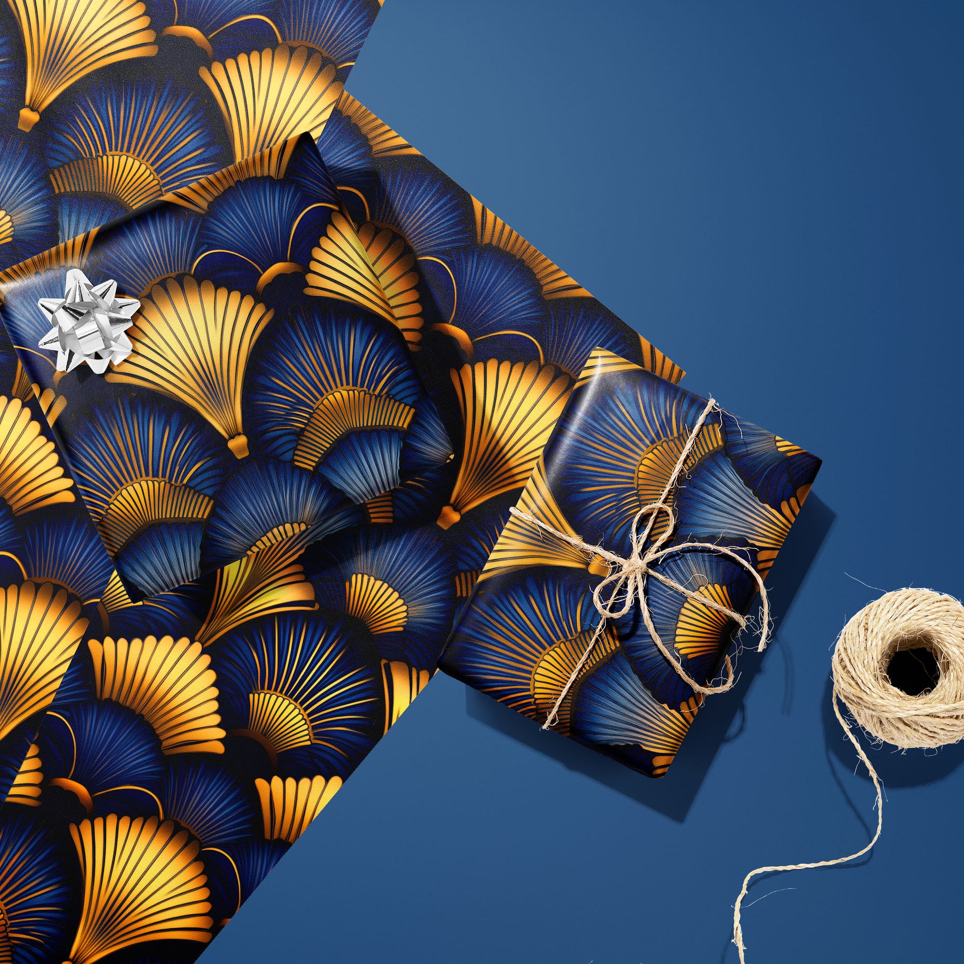 Two gifts in Gilded Ginko—one silver-bowed and one tied with twine—rest on two unused sheets with gold and blue ginkgo leaves, beside a twine spool on a blue surface.