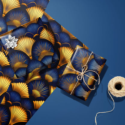 Two gifts in Gilded Ginko—one silver-bowed and one tied with twine—rest on two unused sheets with gold and blue ginkgo leaves, beside a twine spool on a blue surface.
