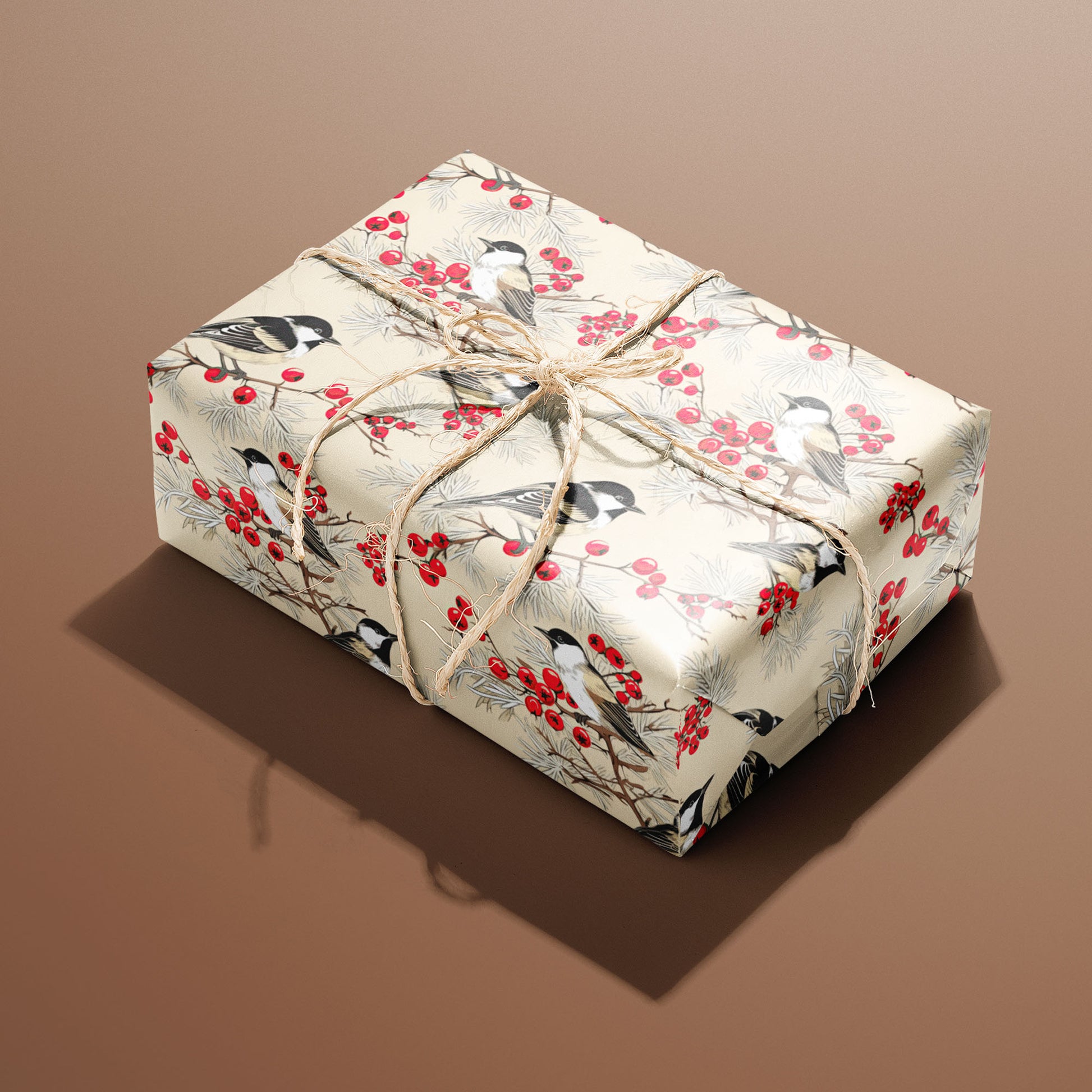 A rectangular gift wrapped in Bird & Berry paper and tied with natural twine displays chickadees among red berries and pine on a cream ground against a warm brown background.