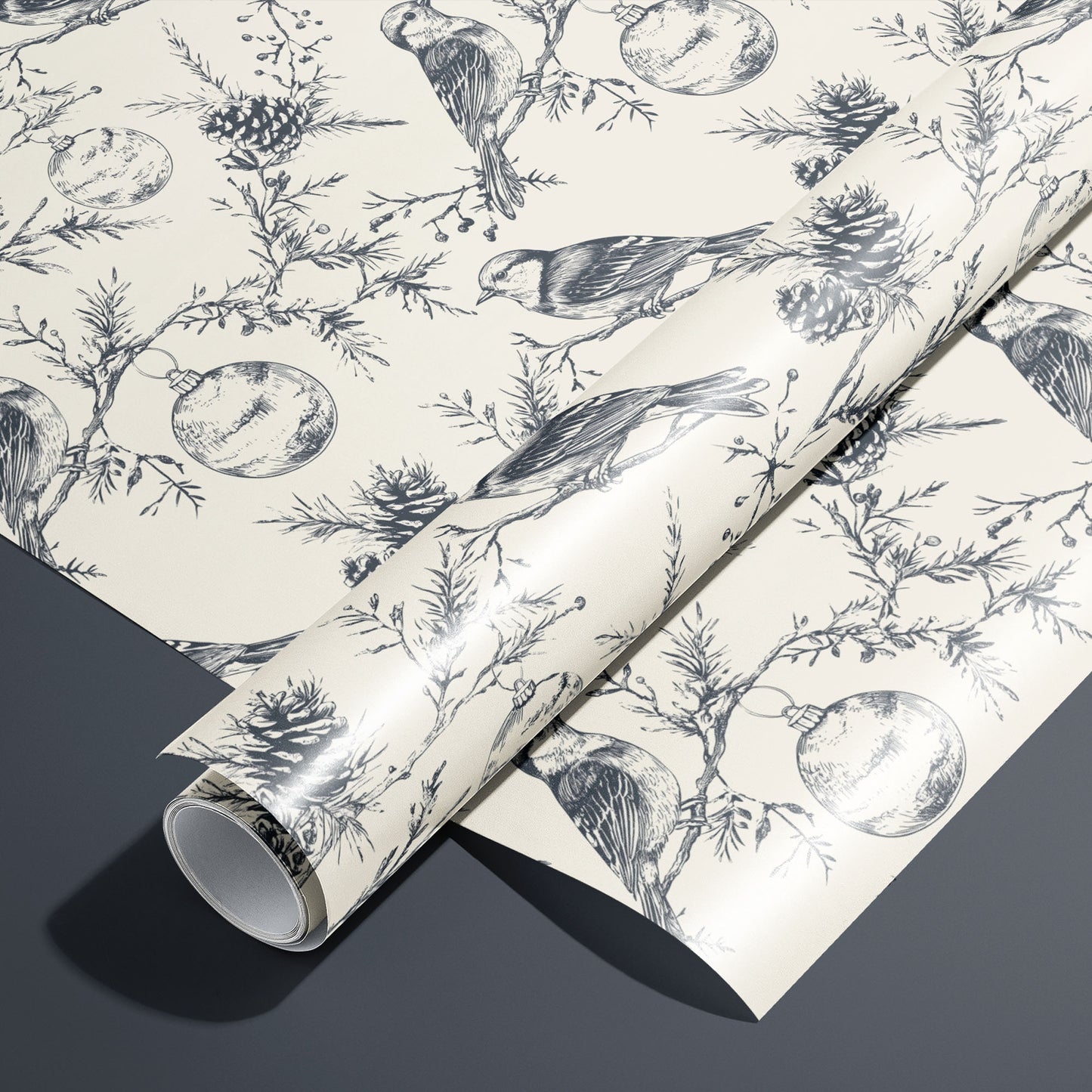 A roll of Winter Wings wrapping paper on an unrolled sheet, featuring charcoal sketch songbirds, pinecones, and ornaments on a cream ground, set on a dark grey surface.