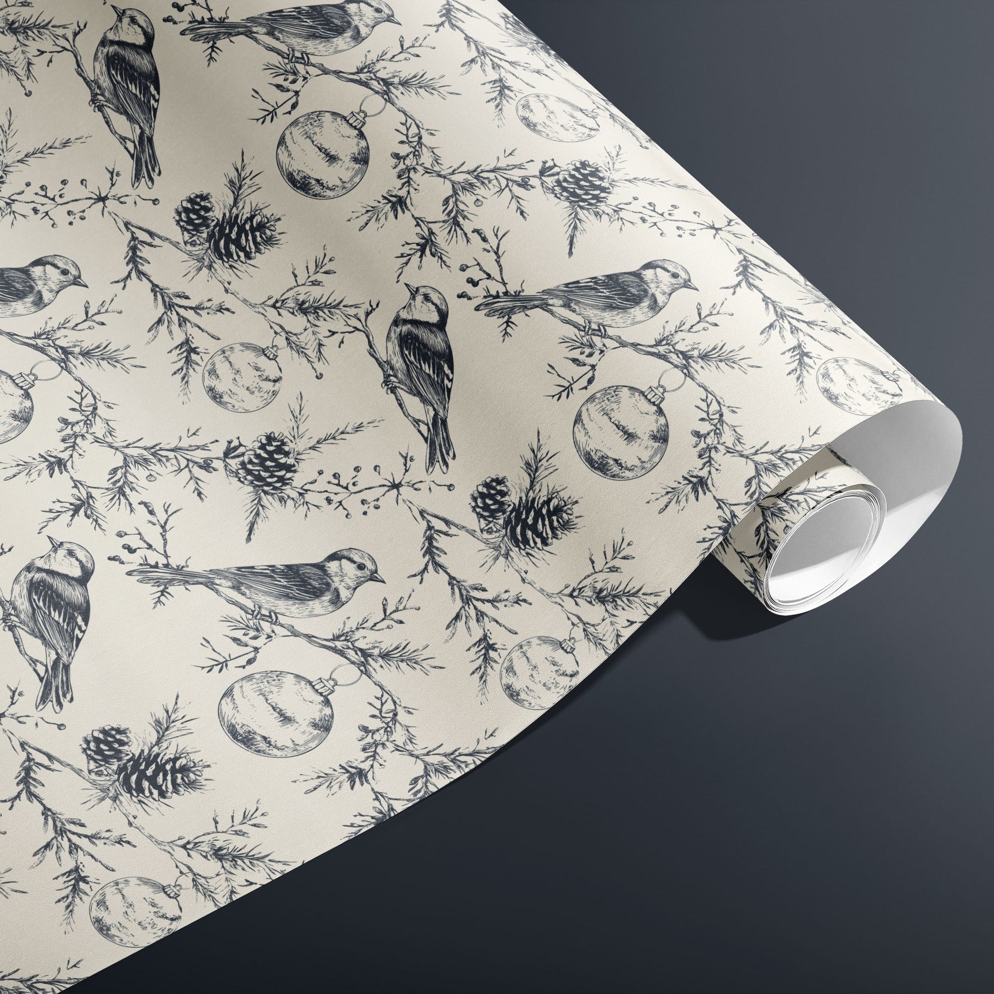 A partially unfurled roll of Winter Wings wrapping paper with hand-drawn birds, pine sprigs, and pinecones in charcoal on cream, resting on a dark grey surface.
