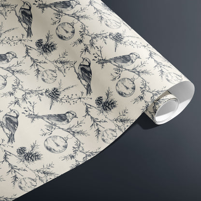 A partially unfurled roll of Winter Wings wrapping paper with hand-drawn birds, pine sprigs, and pinecones in charcoal on cream, resting on a dark grey surface.