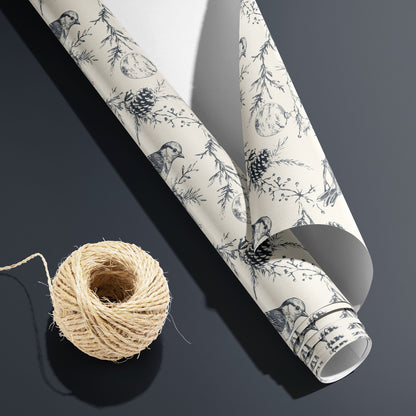 A partially unfurled roll of Winter Wings showing its white underside beside a spool of natural twine, with charcoal sketch birds and pine boughs on cream over a dark grey surface.