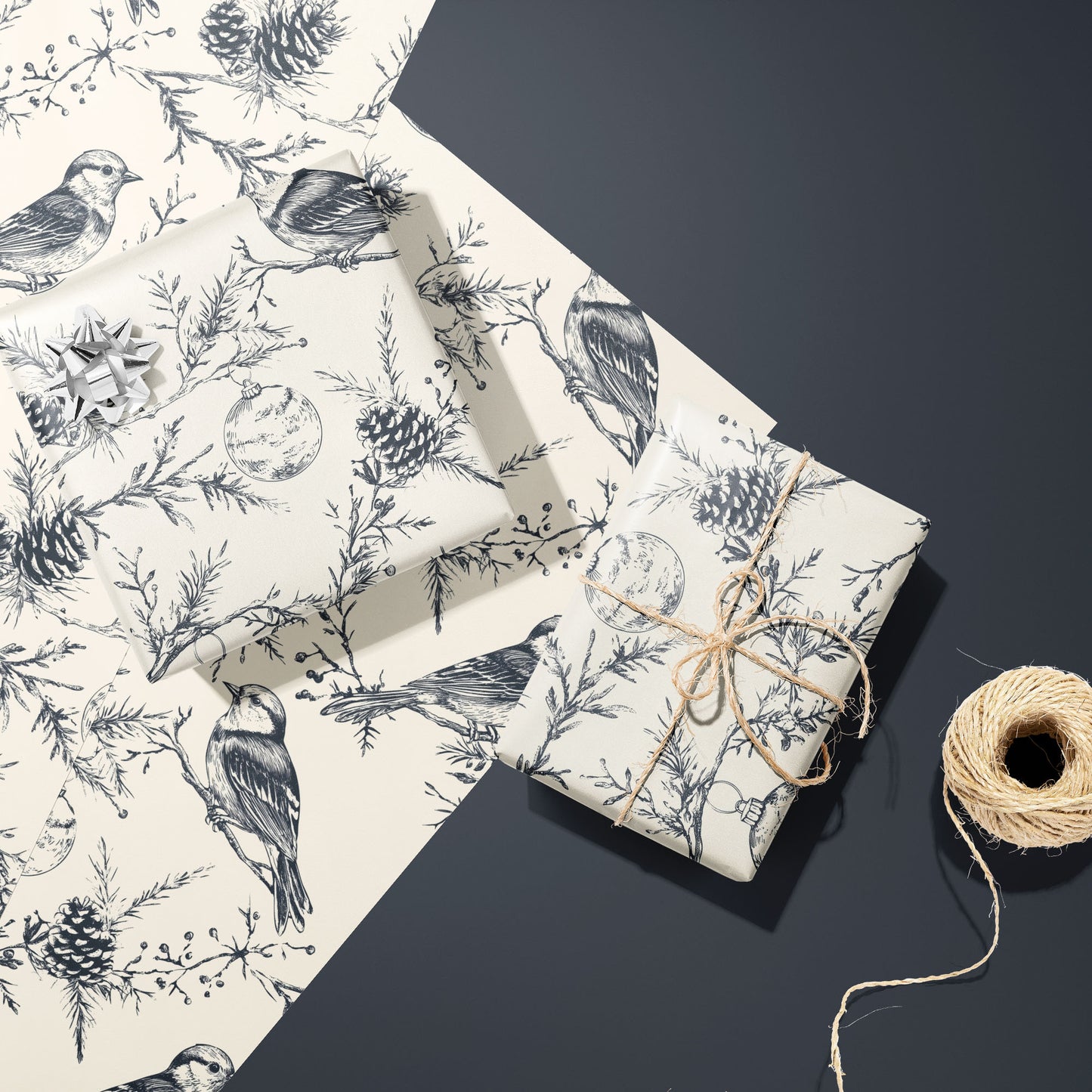 Two gifts wrapped in Winter Wings—one with a silver bow, one tied with twine—rest on two unused sheets beside a twine spool on a dark grey surface, with birds and pine motifs.