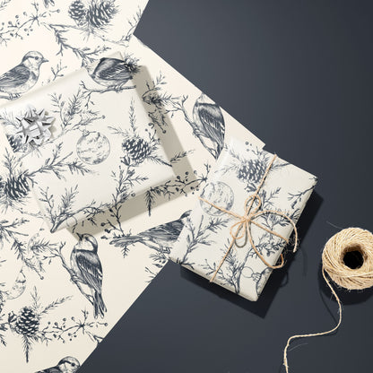 Two gifts wrapped in Winter Wings—one with a silver bow, one tied with twine—rest on two unused sheets beside a twine spool on a dark grey surface, with birds and pine motifs.