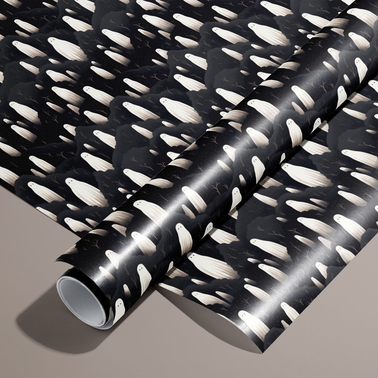 A roll on an unrolled sheet of Halloween Ghosts wrapping paper with ivory ghosts drifting through charcoal clouds and twiggy branches on black, set on a warm taupe background.