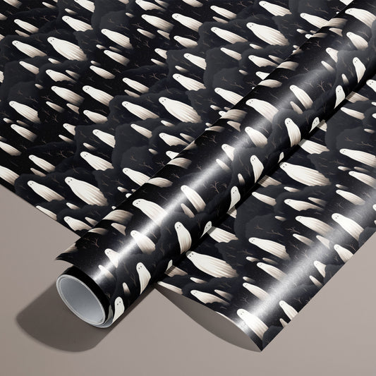A roll on an unrolled sheet of Halloween Ghosts wrapping paper with ivory ghosts drifting through charcoal clouds and twiggy branches on black, set on a warm taupe background.