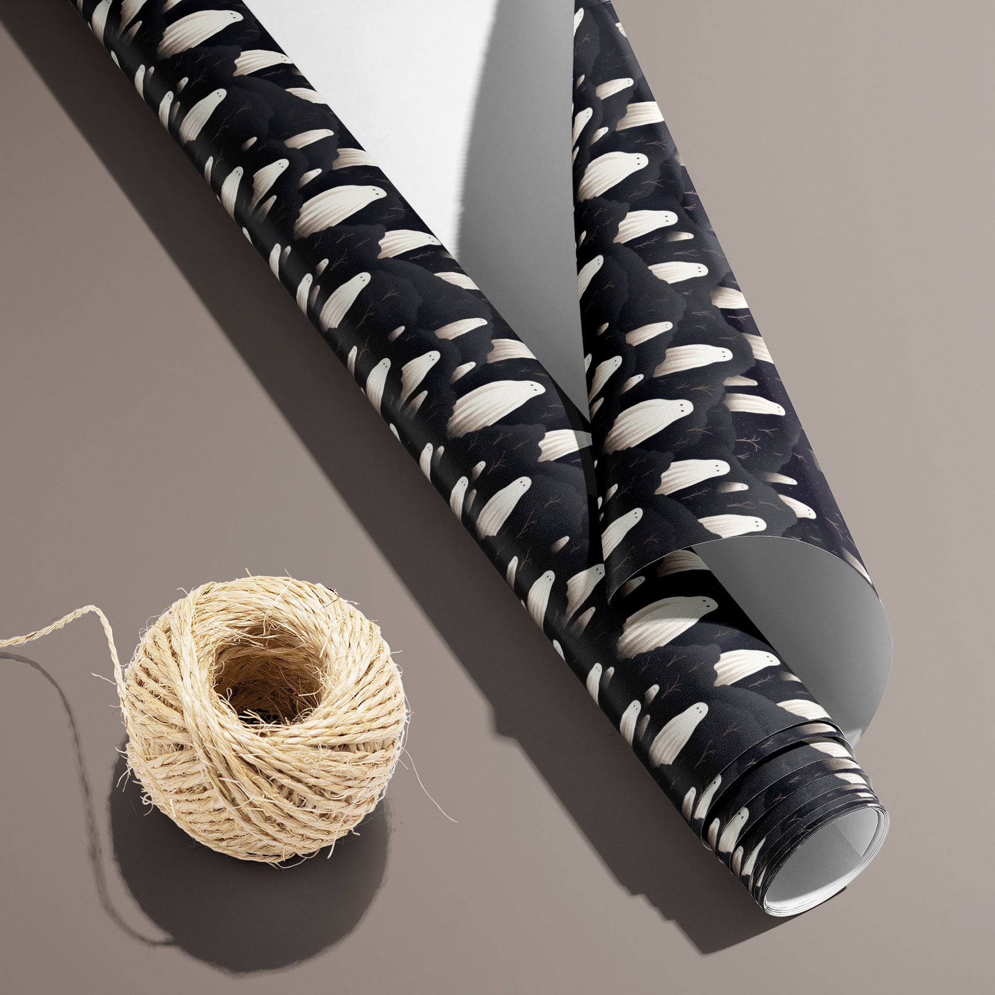 A partially unfurled Halloween Ghosts roll with the white underside visible beside a spool of natural twine, featuring ivory ghosts on black on a warm taupe surface.