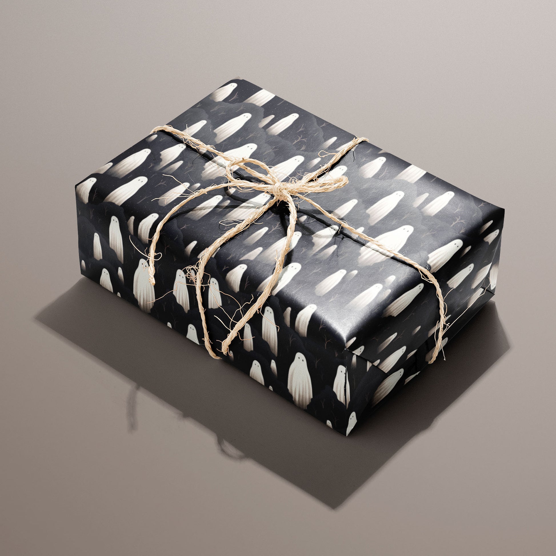 A single rectangular gift wrapped in Halloween Ghosts paper—ivory spirits amid charcoal clouds on black—tied with natural twine on a warm taupe background.