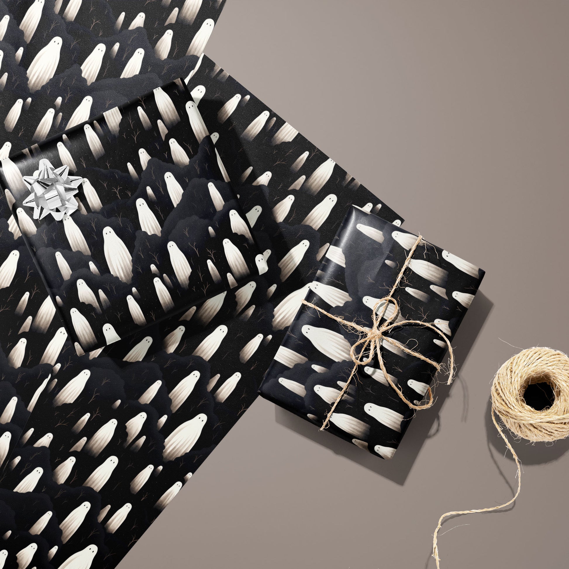 Two gifts in Halloween Ghosts wrap—one with a silver bow and one tied with twine—rest on two unused sheets beside a spool of twine on a warm taupe background.